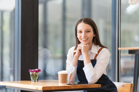 Portrait of happy small business owner sitting at counter in coffee shop. asian female barista wearing apron smiling at bar in cafe. food service, restaurant concept.の写真素材