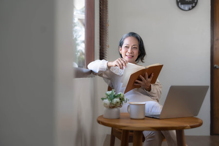 Relaxed and calm 60s retired Asian woman looking out the window, daydreaming while reading a book in her home living room. lifestyle conceptの写真素材