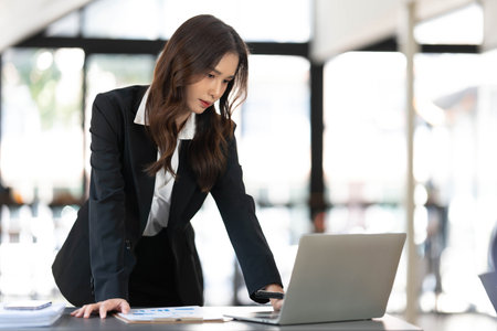 Focused young businesswoman standing at table in office, using laptop, looking at computer screen, reading or writing business email, searching information in internet, working on projectの写真素材