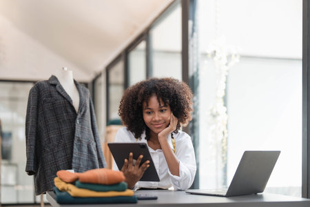 Portrait of young female tailor with tape measure in her neck sitting in stylish dress and holding digital tablet in officeの写真素材
