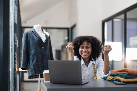 Millennial Asian young professional female designer dressmaker businesswoman with measuring tape on neck sitting smiling hold fists up celebrating together in tailor workshopの写真素材