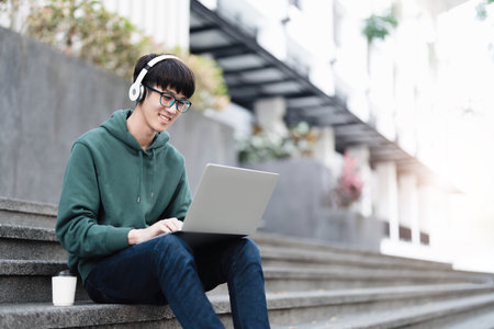 Smart Asian male college student wearing headphones, using laptop on campus outdoor stairsの写真素材