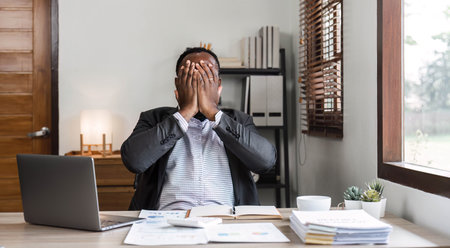 Stressed business man sitting at office workplace. Tired and overworked black man. Young african american exhausted men in stress working on laptop computerの写真素材
