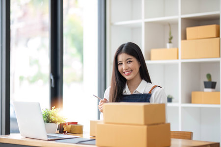 A portrait of a young Asian woman, e-commerce employee sitting in the office full of packages in the background write note of orders and a calculator, for SME business ecommerce and delivery businessの写真素材