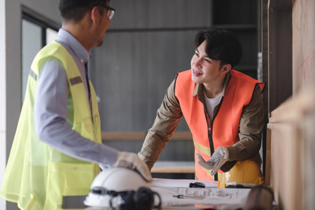 young man construction team engineer in vest and hat working with laptop standing on construction site House building project. Engineer talking with colleagues at workの写真素材