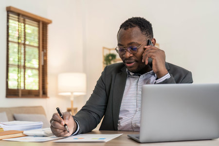 Portrait of handsome African black young business man working on laptop and talking phone at office desk.の写真素材