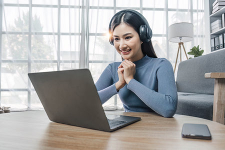 Smiling young asian woman using laptop web camera while wearing headphones and sitting on the rug beside to the sofa at homesの写真素材