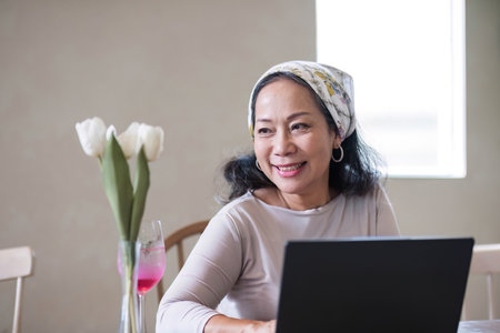 Attractive beautiful mature Asian woman sits at a table with her use laptop in her minimal living room. Hobby and lifestyle conceptsの写真素材