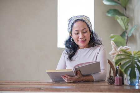 Attractive beautiful mature Asian woman sits at a table with her use laptop in her minimal living room. Hobby and lifestyle conceptsの写真素材