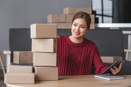 A portrait of a young Asian woman, e-commerce employee sitting in the office full of packages in the...