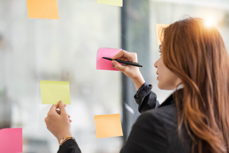 Close-up image of a beautiful young Asian female marketing assistant looking at the sticky note on a glass wall. ideas, notes, memo boardの写真素材