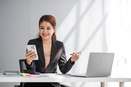 Attractive brunette Asian woman using mobile phone, smile and happy while sitting at working desk in modern office.の写真素材