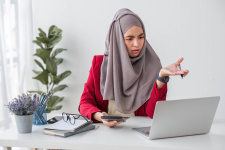 Serious young woman wearing hijab sitting at desk working on new business project in modern officeの写真素材