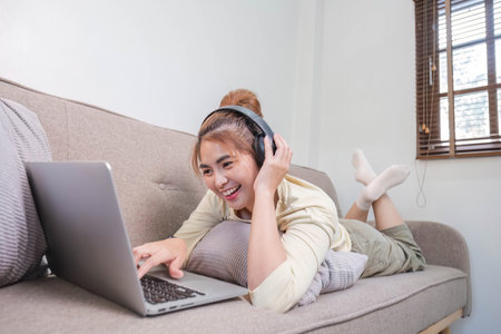 Young beautiful asian woman relaxing and listening to music using headphones, she is lying in sofa. Young woman enjoying at home using laptop.の写真素材