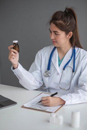 Young beautiful asian woman doctor holding pills bottle writing on document at clinicの写真素材