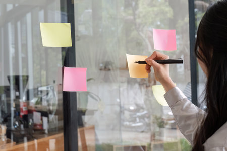 Close up hand businesswoman writing sticky notes on glass wall in officeの写真素材