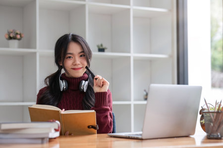 Asian female student studying online using headphones and laptop taking notes in notebook sitting at...
