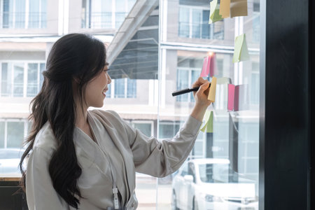 Close up hand businesswoman writing sticky notes on glass wall in officeの写真素材