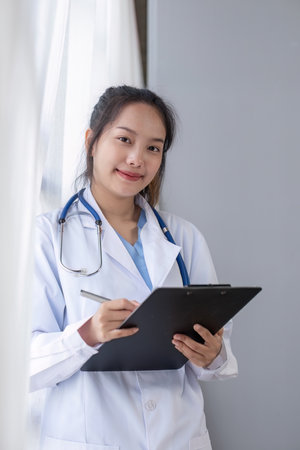 Portrait of female doctor wearing white coat with stethoscope holding clipboard and looking confidently to cameraの写真素材