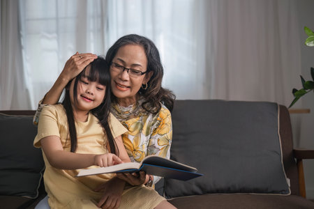Grandma and granddaughter happily spend their free time reading books in the living room. An expression of love between an elderly woman and her adorable granddaughter.の写真素材