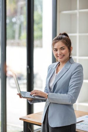 Portrait, Professional and confident Asian businesswoman or female executive manager in formal suit and use laptop. standing, leaning on table, holding laptop and using laptop computer.の写真素材