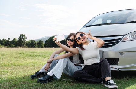 Two young women in white t-shirts and jeans sit back and relax, taking selfies together with their cell phones.の写真素材
