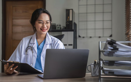 Portrait of cheerful smiling young female doctor in blue medical uniform typing on laptop computer, sitting at desk near window in modern office of medic clinicの写真素材
