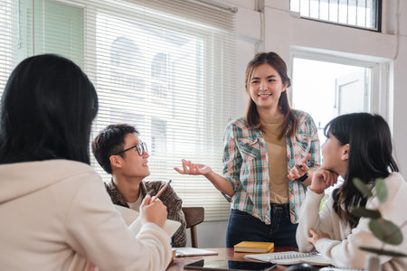 A cheerful and smart young Asian female is standing and sharing her ideas in a meeting with her team. University students, friendship, startups, teamworkの写真素材