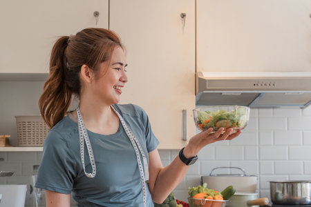 Smiling fitness nutritionist in casual clothes On the table with a laptop computer Holding a bowl of organic vegetable salad Place a measuring tape around your neck in a modern kitchen at homeの写真素材