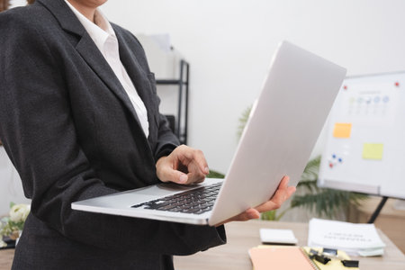 Close up, Young Asian businesswoman with a bright smile holds a laptop and stands in front of a work...