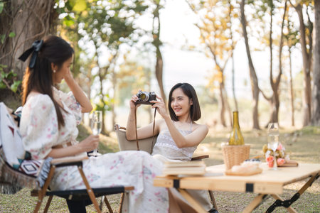 Two beautiful LGBT young women in casual clothes and summer hats Carefree woman having a picnic outside Positive model sitting on the grass eat fruit and cheese Take a selfie. LGBT conceptの写真素材