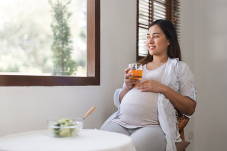 A happy pregnant woman sit, relax, drink water and eats a healthy salad on the dining room table in the living room.の写真素材