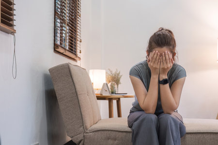 Unhappy anxious Asian woman sitting covering her face with stress in living room at homeの写真素材