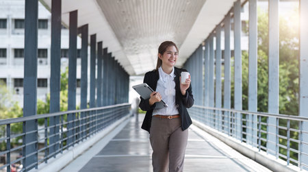 Asian businesswoman holding a laptop and a coffee mug. Smiling at the camera. Confident female employee holding a laptop standing in front of the camera on the background of a city skywalk.の写真素材
