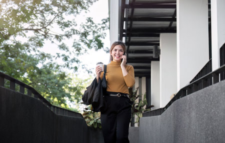 Asian businesswoman smiling while talking on mobile phone and holding coffee cup outside the office.の写真素材