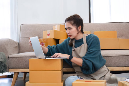 Young female small business entrepreneur dealing with online sales is stressed using her laptop to check product orders and prepare them for delivery.の写真素材