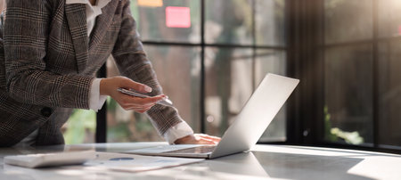 Smiling Asian businesswoman uses laptop to work on financial transactions, tax calculations, planning at work table in glass office.の写真素材