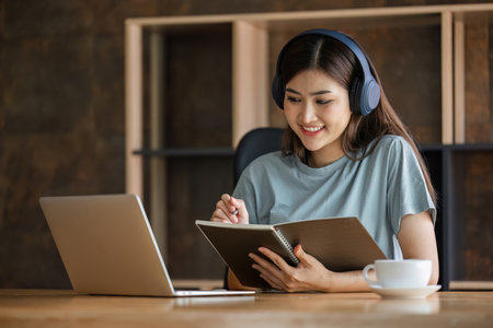 Young female student wearing headphones sits intently and happily studying online on her laptop in the living room at home.の写真素材