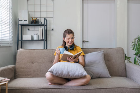 Young Asian woman sitting on modern sofa in front of window, relaxing in living room, reading a book at home.の写真素材