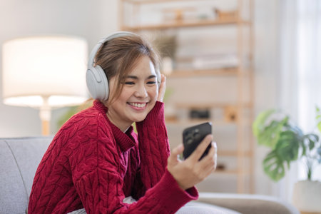 Asian woman sitting on sofa in living room using smartphone and listening to music on headphones.