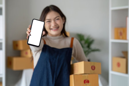 Beautiful smiling Asian woman holding a cardboard box and hands showing a smartphone mockup of a blank screen. Advertise your screen Online delivery conceptの写真素材