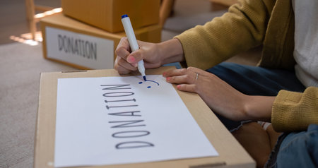 A beautiful Asian woman uses a magic marker to write on a box of clothes that will be donated to...