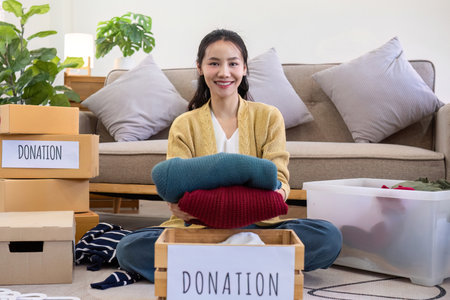 Cute Asian woman sitting next to sofa in living room sorting unwanted clothes for donation.