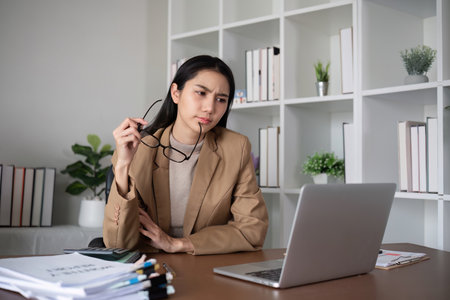 Young Asian business woman sits and works using a laptop in a modern office decorated with shady green plants.の写真素材