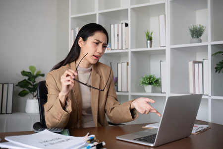 Unhappy Asian business woman shows stress over unsuccessful business while working in home office decorated with soothing green plants.の写真素材