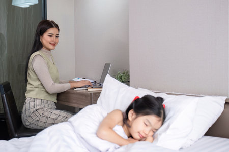 Young woman, single mother, with cute daughter in bedroom A young woman is working online on her laptop from home with her daughter waiting on her bed.の写真素材