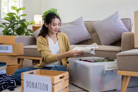 A young Asian woman sits on a sofa sorting unwanted clothes to donate to a charity.