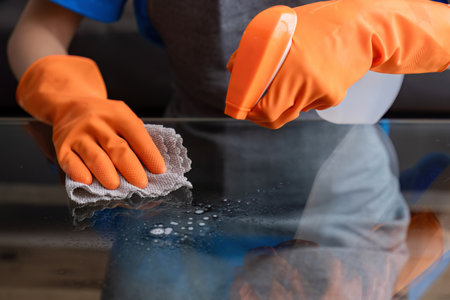 Beautiful young housewife wearing orange housekeeping gloves uses spray and cloth to clean the table...