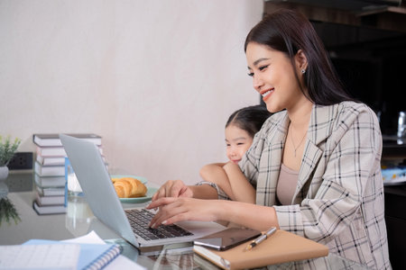 a single mother, sits at home working on a laptop with her daughter beside her watching and encouraging her.の写真素材