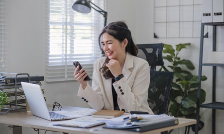 Young Asian business woman sits on the phone in an online business meeting using a laptop in a modern home office decorated with shady green plants.の写真素材